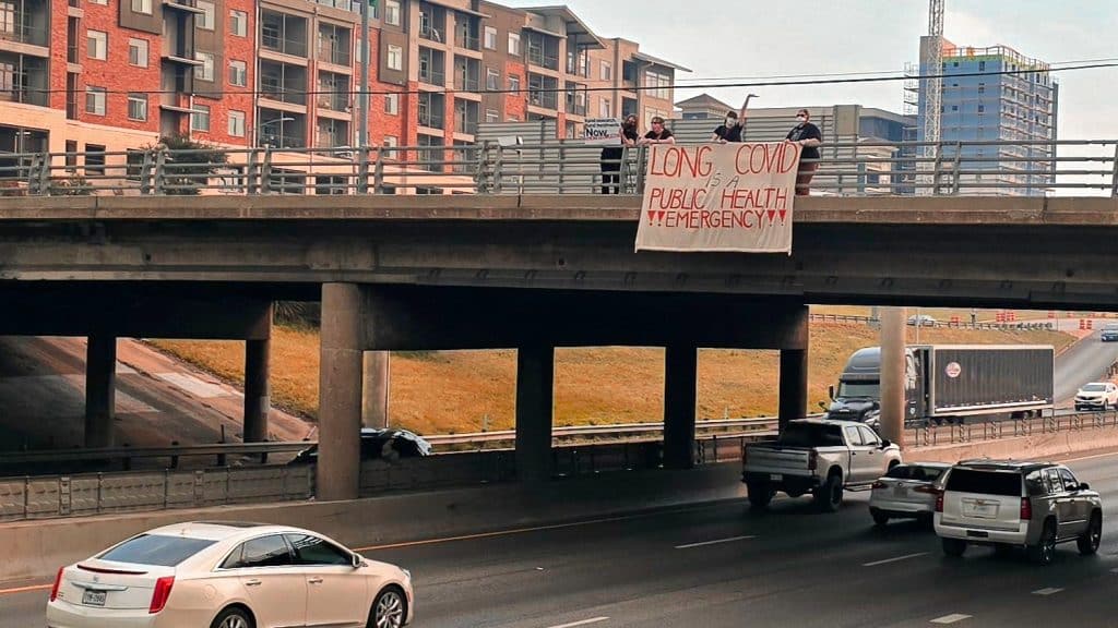 A photo of 3 people masked and holding a banner over the side of an overpass on a highway. The banner has bold red and black text that reads, "Long COVD is a public health emergency!!"