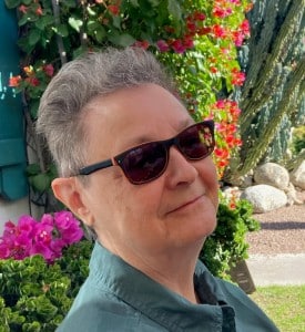 Author photo of Shelley Ettinger, featuring an older woman in dark sunglasses with short grey hair standing in front of backyard flowers.
